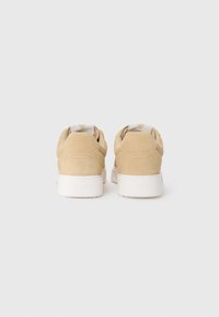 Beige mokkasiner i semsket skinn med en hvit gummisåle og et strukturert bakpanel. Har en stoffbit med en trekantet aksent.