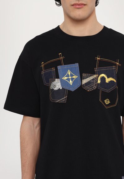 T-shirt noir avec cinq poches décoratives de différentes teintes de denim bleu et de couleurs métalliques, accompagnées de détails de couture et de designs uniques.