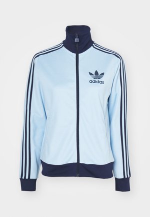 Lyseblå treningsjakke med marineblå krage og mansjetter, med tre marineblå striper på ermene og et Adidas-logo. Glidelås-lukking, glatt stoff.