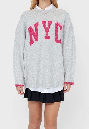 Mujer con un suéter gris claro que lleva grandes letras rosas "NYC", una camisa blanca debajo y una falda negra plisada.
