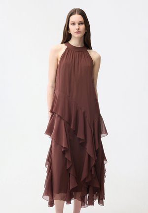 Frau trägt ein ärmelloses braunes Kleid mit hohem Kragen und geschichteten, asymmetrischen Rüschen auf weißem Hintergrund.
