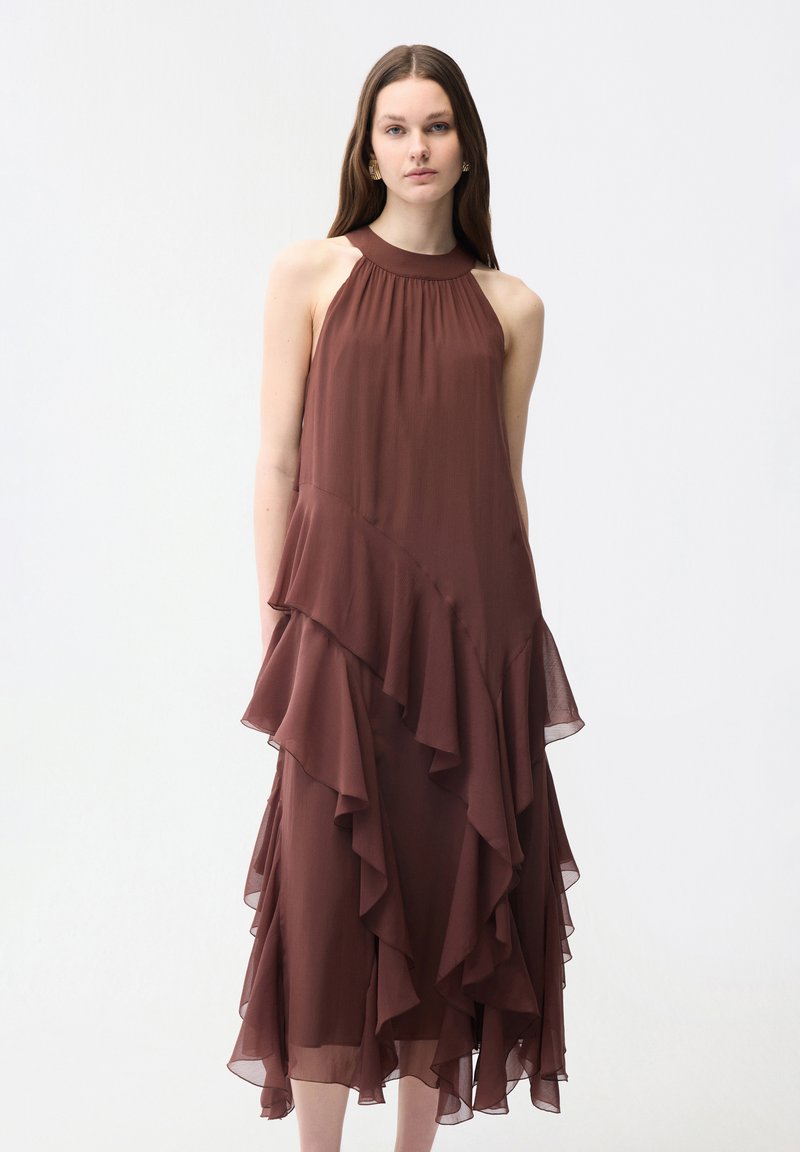 Frau trägt ein ärmelloses braunes Kleid mit hohem Kragen und geschichteten, asymmetrischen Rüschen auf weißem Hintergrund.