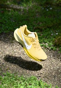 Baskets de sport jaunes avec une tige texturée, un swoosh blanc et une semelle en caoutchouc. Volent au-dessus du gravier et de la verdure. Design léger.