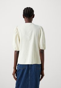 Top con maniche a palloncino in tessuto crema chiaro, con collo rotondo e una texture liscia. Indossato con una gonna in denim scuro, che mostra dettagli di design sottili.