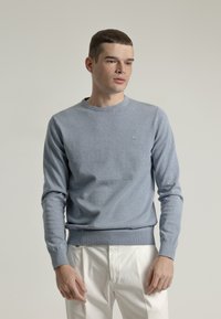 Suéter de punto azul claro con mangas largas y cuello redondo, con un pequeño logotipo en el pecho y un dobladillo acanalado.