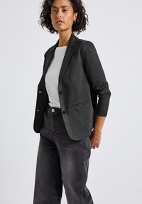 Blezer w kolorze węgla drzewnego o dopasowanym kroju, z zapięciem na jeden guzik oraz dwiema przednimi kieszeniami. Noszony na białej koszuli i zestawiony z szarymi dżinsami.