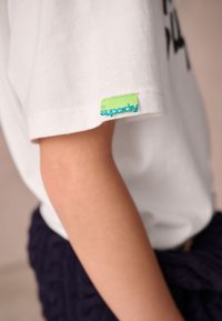 Närbild på en persons arm som bär en vit t-shirt med en liten grön tagg märkt "by superdry" nära ärmsluten.