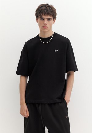 Ung mand med kort brunt hår iført sort oversized T-shirt og bukser, står mod en ensfarvet hvid baggrund med den ene hånd i lommen.