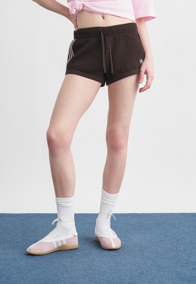 Person trägt schwarze, strukturierte Shorts mit Seitenstreifen, ein gebundenes rosa Crop-Top, weiße Socken und rosa Schnürschuhe und steht auf blauem Teppich.