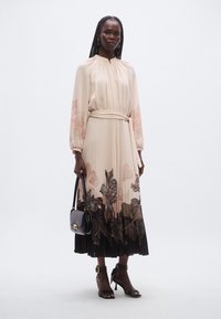 Lysebeige kjole med blomsterprint i mørke toner, plisseret nederdel, lange ærmer og bælte. Stylet med en lille sort håndtaske og hælede sandaler.