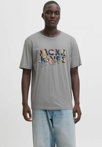 Pelēka kokvilnas t-kreklu ar daudzkrāsainu "JACK & JONES" logo apdruku. Īsas piedurknes, apaļš kakls, brīva piegriezuma. Nēsāts ar gaiši ziliem džinsiem.