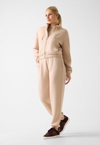 Jasno beżowa bluza na zamek z wysokim kołnierzem, dopasowane dresowe spodnie. Brązowe sneakersy z białymi podeszwami. Prosty design, miękki materiał.