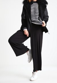 Veste en similicuir noir avec col en fourrure, pull en tricot à motif, pantalon noir ample et baskets montantes blanches.