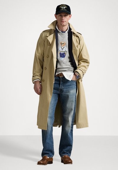 Beige trench coat over a gray sweater with an illustration, blue jeans, and brown shoes. Black cap with yellow text.