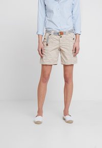 Beige shorts dengan saku depan, kain bertekstur, dan ikat pinggang anyaman yang dapat disesuaikan dengan garis biru dan putih. Dipakai dengan espadrilles putih.