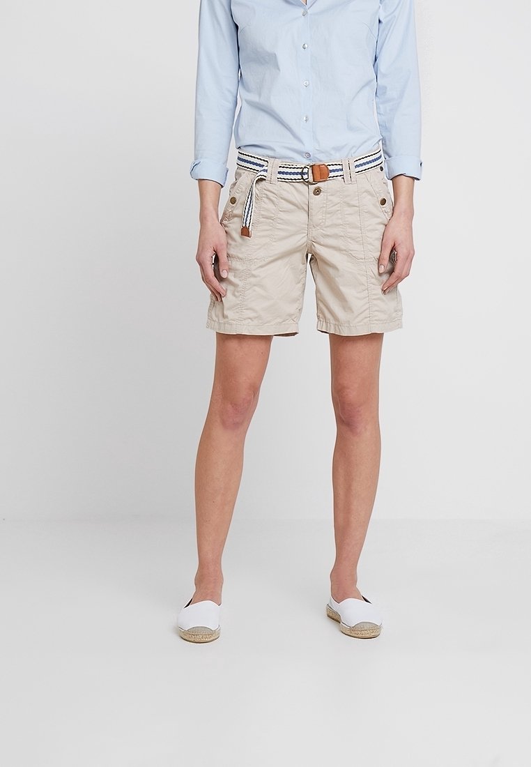 Beige shorts dengan saku depan, kain bertekstur, dan ikat pinggang anyaman yang dapat disesuaikan dengan garis biru dan putih. Dipakai dengan espadrilles putih.