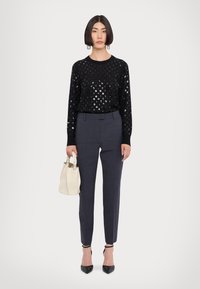 Schwarzer Pullover mit silbernen Pünktchen, kombiniert mit grauen maßgeschneiderten Hosen. Die Accessoires umfassen eine kleine weiße Handtasche und schwarze spitze Absätze.