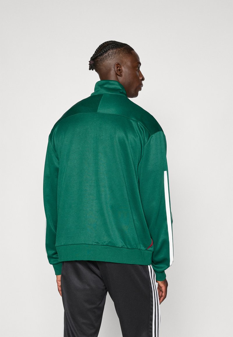 Groene sportjas met hoge kraag, gestructureerde stof en witte zijstrepen. Draag met zwarte broek met drie witte strepen.