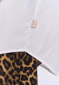 Biała koszula o subtelnej teksturze, z dołem wykończonym beżową metką; połączona z brązowym i czarnym materiałem w panterkę poniżej.