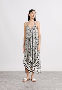 Μακρύ φόρεμα με ασπρόμαυρο μοτίβο, με λαιμόκοψη halter, ρέουσα σιλουέτα και ασύμμετρο τελείωμα, συνδυασμένο με λευκά σανδάλια slide.