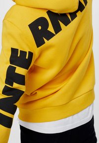 Żółta bluza z dużymi czarnymi literami "INTERN" na rękawie. Posiada kaptur ze sznurkiem i ściągacze na mankietach. Miękka w dotyku.