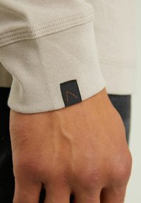 Beige long-sleeve shirt cuff with ribbed texture, featuring a dark rectangular label with a distinctive orange logo stitch.