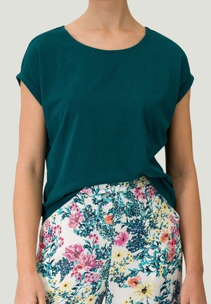 Personne portant une chemise à manches courtes vert foncé rentrée dans un pantalon blanc avec un motif floral coloré.