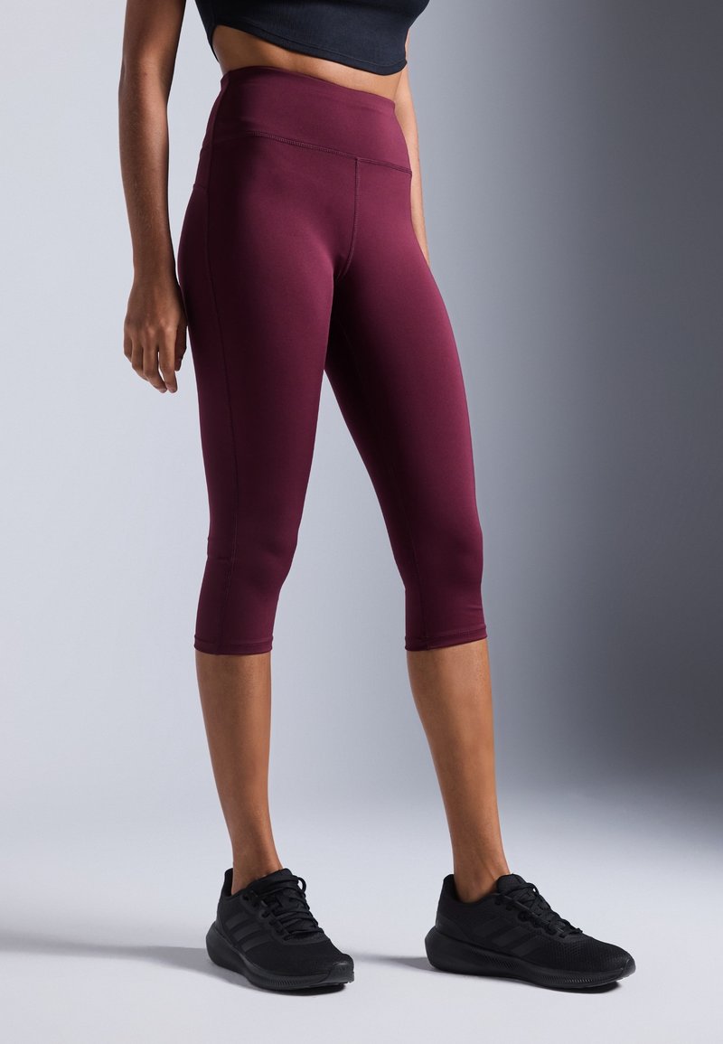 Femme portant des leggings bordeaux taille haute courts et des chaussures de sport noires, debout devant un fond gris uni.