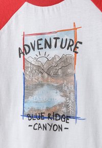 Medvilninis marškinėliai su raudonais raglan rankovėmis, turintis lauko grafiką, įskaitant kalnus, saulę ir tekstą: "Nuotykiai Blue Ridge Canyon."