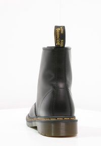 Svart läderankelboot med rund tå, gula sömmar och svart strukturerad sula. Har en gul och svart dragflik på baksidan.