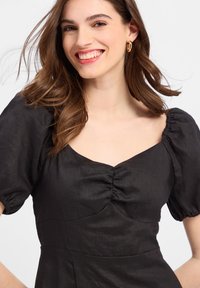 Schwarzes Kleid mit Puffärmeln und einem Herz-Ausschnitt, das vorn Raffung und einen glatten, strukturierten Stoff aufweist. Goldene Ohringe runden den Look ab.