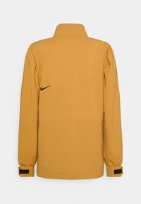 Gul Nike-jacka med hög krage, elastiska ärmslut och svart logotypdetalj på baksidan. Tillverkad av ett mjukt, lätt tyg.