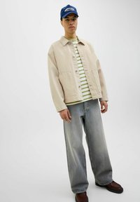 Beige denim jacket over a green and white striped shirt, paired with faded blue baggy jeans and brown shoes. Blue cap completes the outfit.