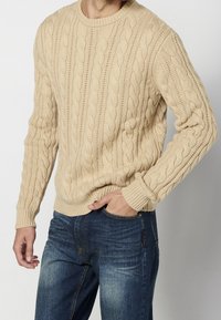 Pull beige en maille torsadée avec encolure ronde et poignets côtelés, associé à un jean bleu foncé. Motifs texturés bien marqués sur toute la surface.