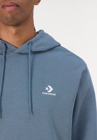 Sudadera azul clara hecha de un tejido suave, con un bolsillo tipo canguro y cordón ajustable. El logo de Converse está bordado en el lado izquierdo del pecho.