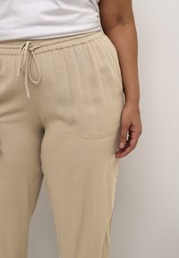 Pantalons beiges légers avec cordon de serrage, à coupe décontractée et poches latérales. Texture lisse avec des détails de couture discrets.