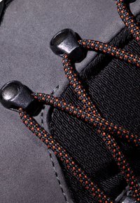 Czarna cholewka buta z teksturowanej tkaniny i czarnymi elementami metalowymi. Sznurowadła mają pleciony wzór z czarnych i pomarańczowych nici.