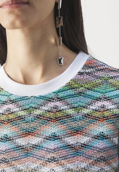 Αντικείμενο μωβ πλεκτή ζακέτα με λευκό ριμπ μανίκι, η οποία διαθέτει σχέδιο ζιγκ-ζαγκ σε μπλε, πράσινους και πορτοκαλί τόνους. Ασημένια γεωμετρικά σκουλαρίκια.