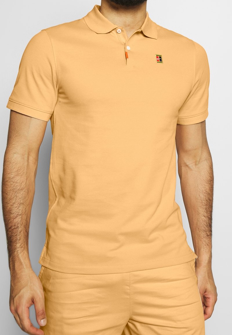 Homme portant un polo à manches courtes orange clair et un short assorti avec un petit logo carré multicolore sur la poitrine.