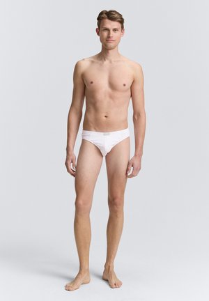 Weiße Baumwollunterhosen mit mittelhohem Schnitt. Glatte Textur, elastischer Bund und minimale Nähdetails. Das Modell steht barfuß aufrecht.