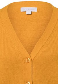 Mosterdgeel gebreide cardigan met een V-hals, geribbelde afwerking en drie gouden knopen aan de voorkant. Zacht textuur en een gedemonteerd wafelpatroon.