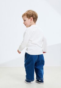 Tricou alb cu mâneci lungi și guler rotund, asortat cu pantaloni din denim cu dungi albastre și adidași închiși la culoare. Copilul este prezentat din spate.