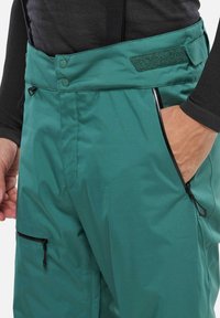 Tropfeneisenfarbene Skihosen verfügen über einen hohen Taillenbund mit Knöpfen, einen verstellbaren Riemen und mehrere mit Reißverschluss versehene Taschen, gefertigt aus einem glatten, strapazierfähigen Stoff.