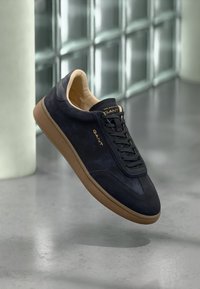 Námornícka semišová teniska s hnedej gumenej podrážkou, s tmavými šnúrkami a zlatým logom. Hladká textúra, nízky profil a zaoblená špička.