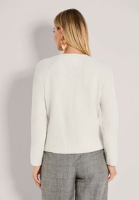 Vrouw met lang blond haar, gekleed in een witte gebreide trui en grijs geruite broek, staand tegen een effen lichte achtergrond en met haar gezicht van zich af.