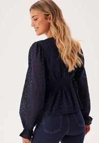 Marineblauwe blouse met volumineuze doorschijnende mouwen, een gesmokt middel en een gaatjespatroon, gecombineerd met donkere denim jeans.