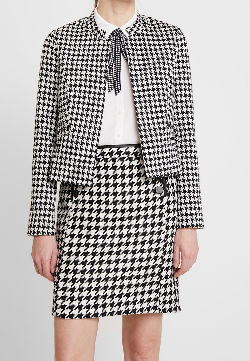Femme portant une veste et une jupe en pied-de-poule noir et blanc avec un chemisier blanc et une cravate assortie pied-de-poule.