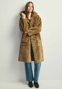 Brązowy płaszcz z sztucznego futra z kapturem, dwiema kieszeniami z przodu i zapięciami na trok. W połączeniu z niebieskimi spodniami dzwonami i ciemnymi botkami za kostkę.