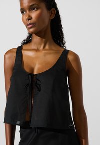 Juodas trumpas viršus, pagamintas iš lengvos medžiagos; turi apvalų iškirpimą, reguliuojamą priekinį raištelį ir minimalistinį dizainą su šiek tiek laisvu siluetu.