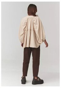 Kobieta w beżowej luźnej bluzce z długim rękawem i marszczonym tyłem oraz brązowych spodniach wąskich, stojąca tyłem do aparatu.